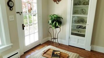 Une pièce avec un plancher en bois, une porte blanche, une fenêtre, une plante posée sur un support en métal, une bibliothèque ornée d’assiettes décoratives et un plateau sur lequel repose un livre intitulé « Le Testament » de John Grisham.