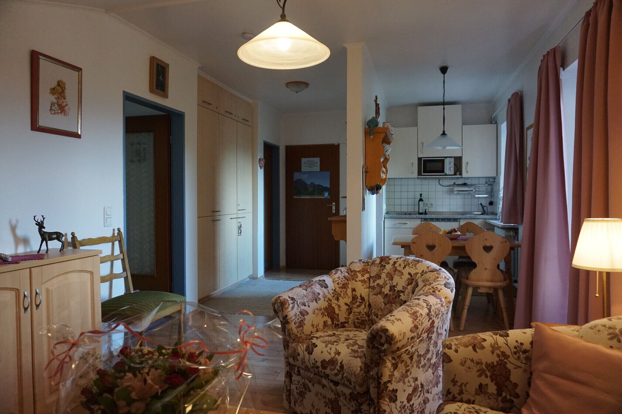 A living room with floral-patterned sofas, a dining table with chairs, a kitchen area with a microwave, and a hallway leading to another room.