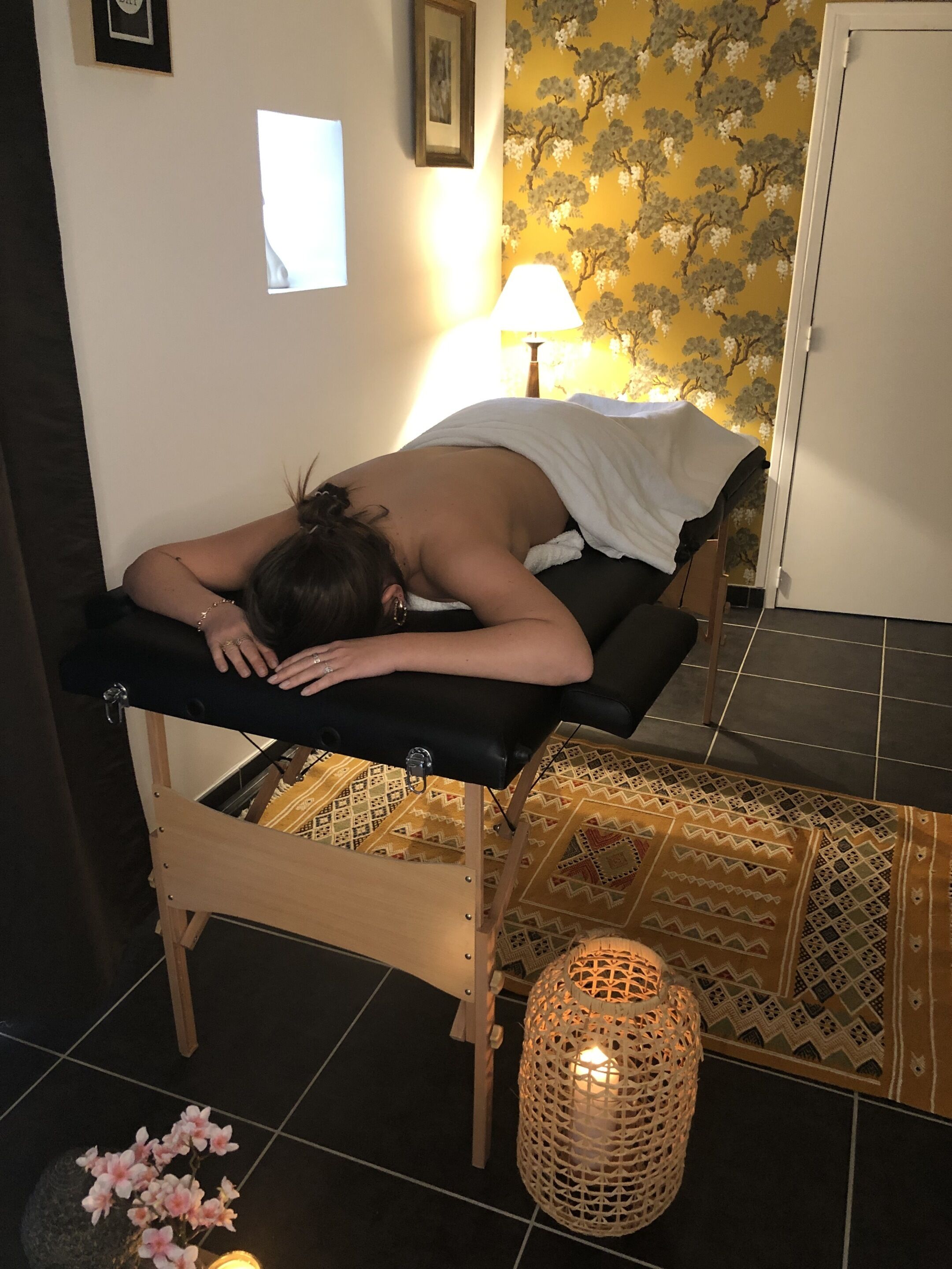 Una persona recibe un masaje sobre una mesa de masaje negra en una habitación con alfombra estampada, una lámpara de pie tejida y una puerta al fondo.