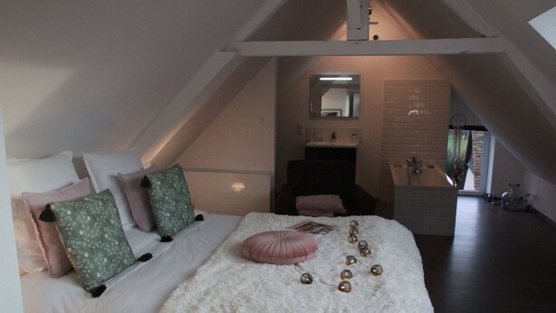 Een slaapkamer met schuine wanden, een bed met wit beddengoed en decoratieve kussens, een pluizig wit tapijt en een kleine badkamer met spiegel en wastafel.
