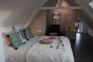 A bedroom with a sloped ceiling, a bed with white bedding and decorative pillows, a fluffy white rug, and a small bathroom with a mirror and sink. - La Catiniere (Arnage)