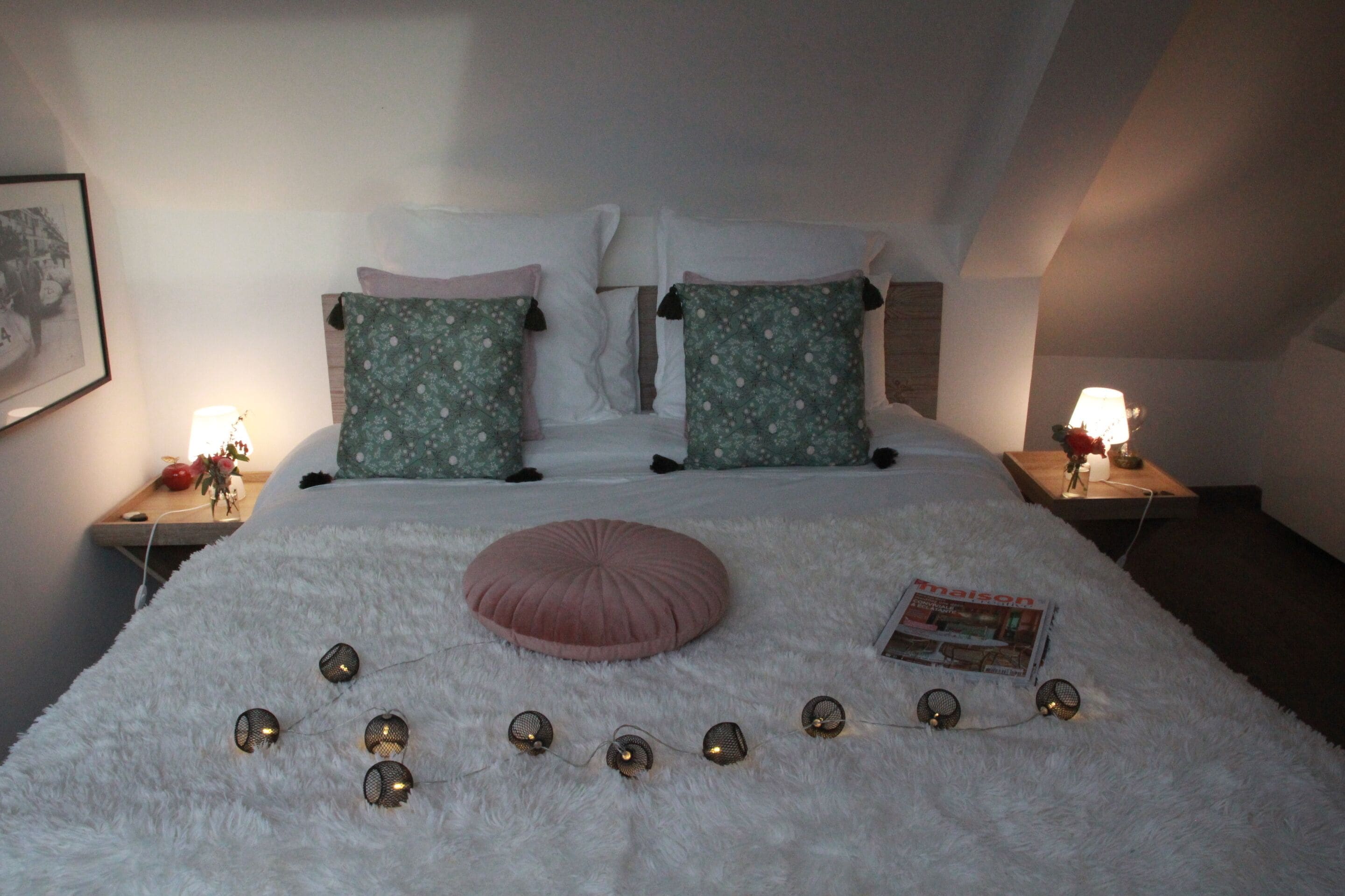 A neatly made bed with a fluffy white cover, two green patterned pillows, a pink round pillow, and a magazine on the bed. There are decorative lights arranged in a circle on the bed.