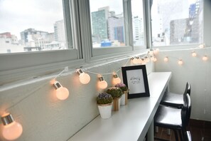 A room with a window overlooking a cityscape, a white table with a picture frame, and a string of warm light bulbs. - Dolma Guesthouse Hongdae (Seoul)