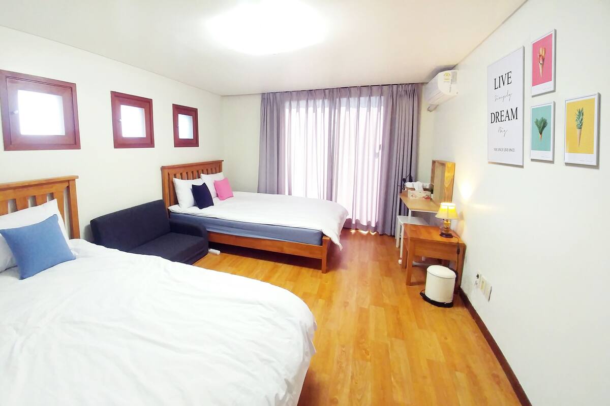 Quarto de hotel com duas camas, uma escrivaninha, uma cadeira, mesa de cabeceira e uma janela com cortinas.