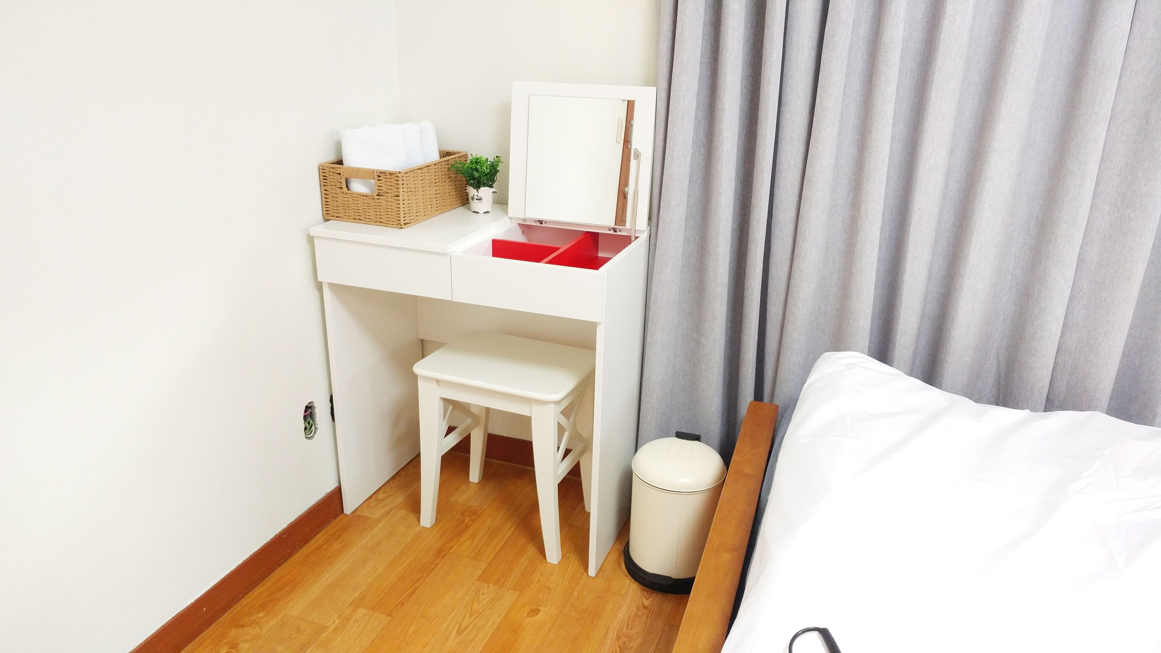 A compact hotel room with a wooden floor, a white desk with a red top, a small white chair, a white bed, and a grey curtain.