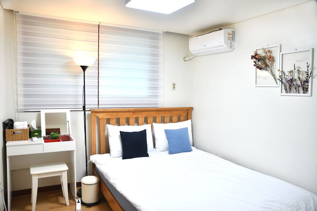 Quarto com cabeceira de madeira, uma cama de solteiro com almofadas brancas e azuis, uma escrivaninha com cadeira, um cesto de lixo e uma janela com persianas.