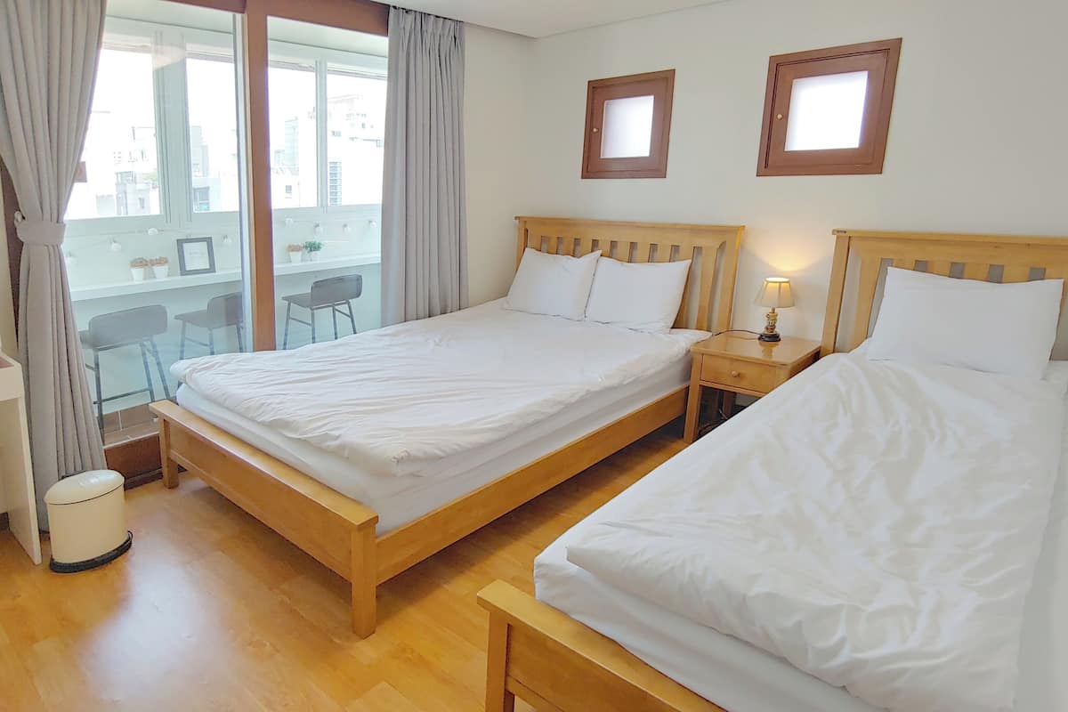 Quarto de hotel com duas camas de madeira, uma pequena mesa de cabeceira com abajur, uma janela com cortinas e vista para a varanda externa.