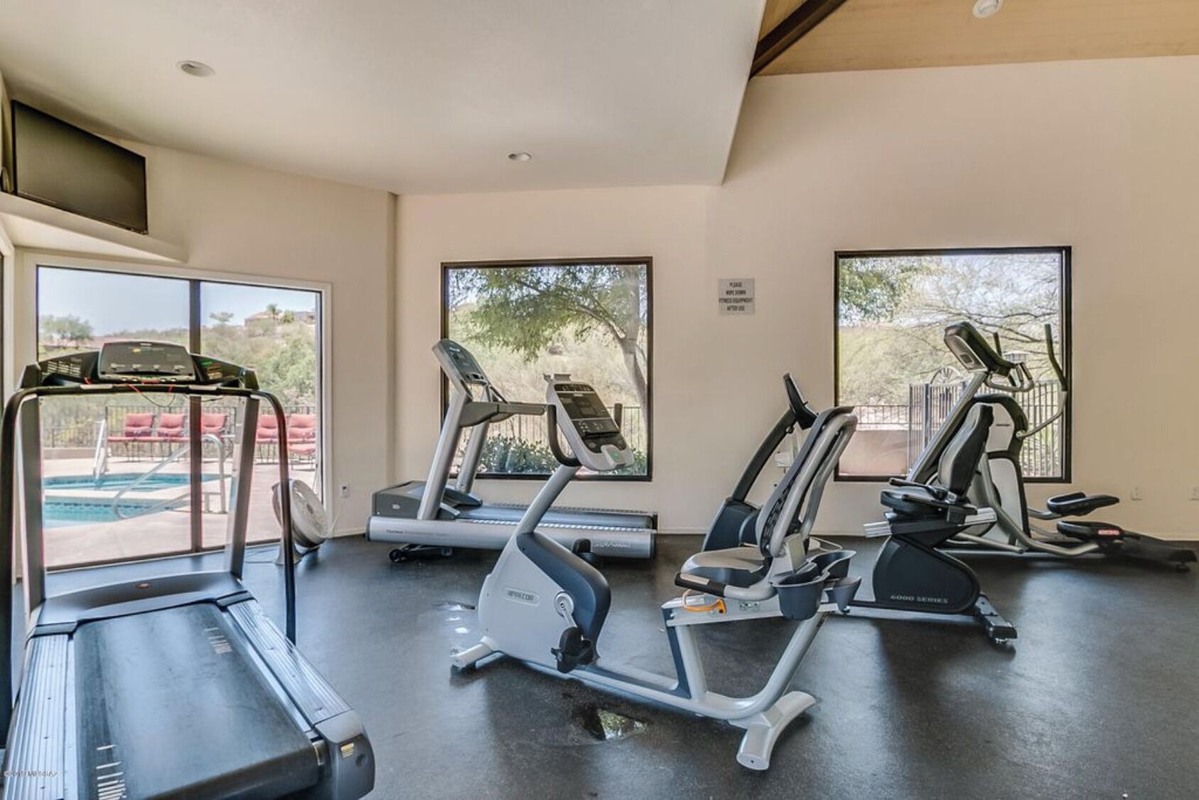 Une salle de sport équipée de tapis de course, de vélos elliptiques et d’une piscine visible à travers les fenêtres.