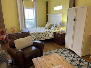 A room with a bed, a brown leather chair, a wooden table, a nightstand with a lamp, and a wardrobe.