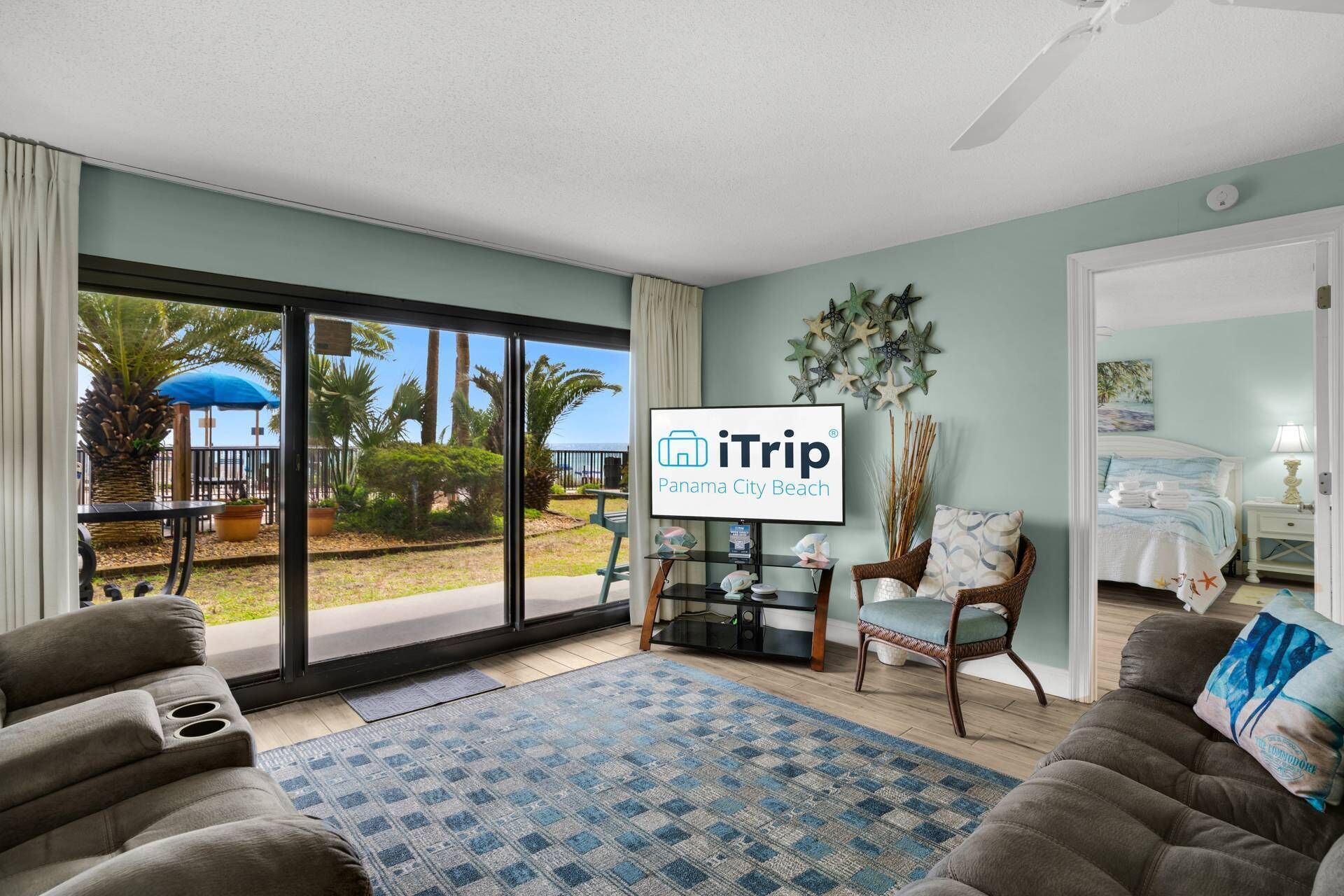 A hotel room with a large sliding glass door leading to a balcony with a view of palm trees and a blue umbrella. There is a flat-screen TV mounted on the wall, a wicker chair with a cushion, and a bed with a blue and white bedding set.