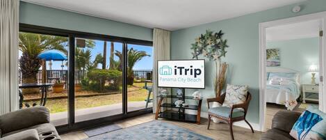 A hotel room with a large sliding glass door leading to a balcony with a view of palm trees and a blue umbrella. There is a flat-screen TV mounted on the wall, a wicker chair with a cushion, and a bed with a blue and white bedding set.