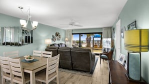 Un salon comprenant un coin repas, un ventilateur de plafond, une grande fenêtre avec vue et un téléviseur posé sur un meuble.