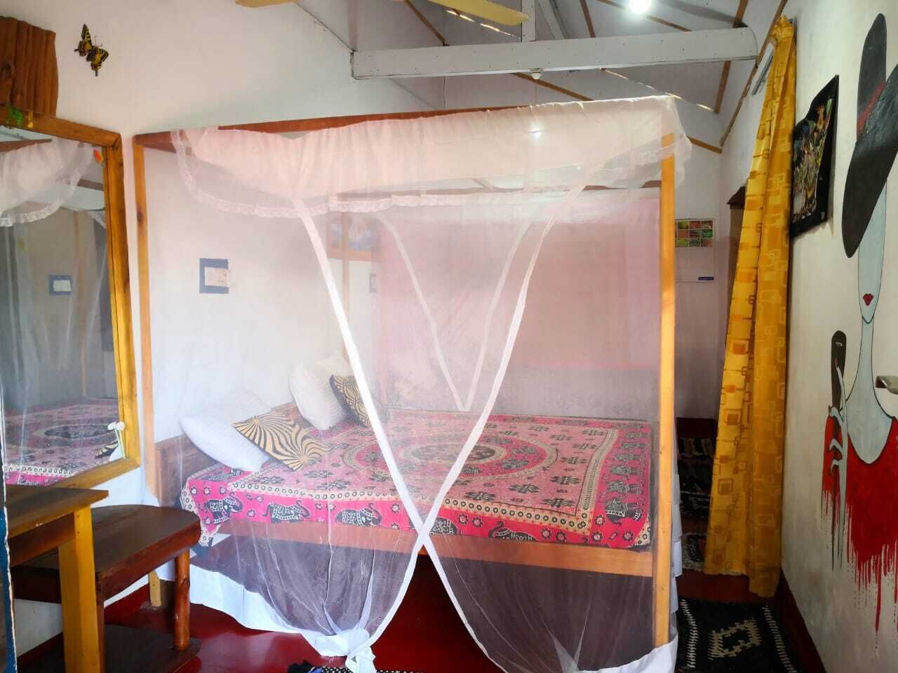 A room with a bed under a mosquito net, a wooden chair, a mirror, and a wall with a painting.