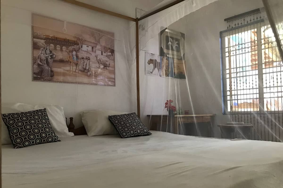 Ein Schlafzimmer mit einem Bett, gemusterten Kissen, einem Moskitonetz und einer Wand mit einem großen Gemälde.
