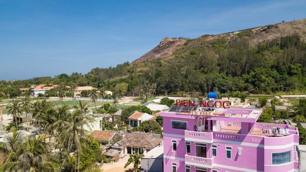 A pink building with a sign reading "BEN NGOC HOTEL CAFE" and a view of a hilly landscape with palm trees and other buildings. - Bien Ngoc Ly Son Hotel (Ly Son)