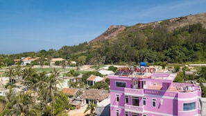 Edifici rosa amb un rètol que diu "BEN NGOC HOTEL CAFE" i una vista d'un paisatge amb turons, palmeres i altres edificis.
