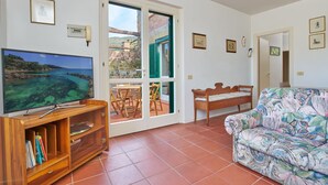 A living room with a television, a wooden cabinet with books, a patterned sofa, and a view of a coastal area through a glass door. - Appartamento Via Dei Lecci - APPARTAMENTO I LECCI (Rio nell'Elba)