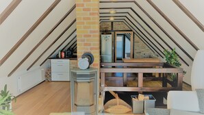 Habitación estilo loft con techo inclinado, chimenea, zona de cocina y área de estar con sofá y cojines.