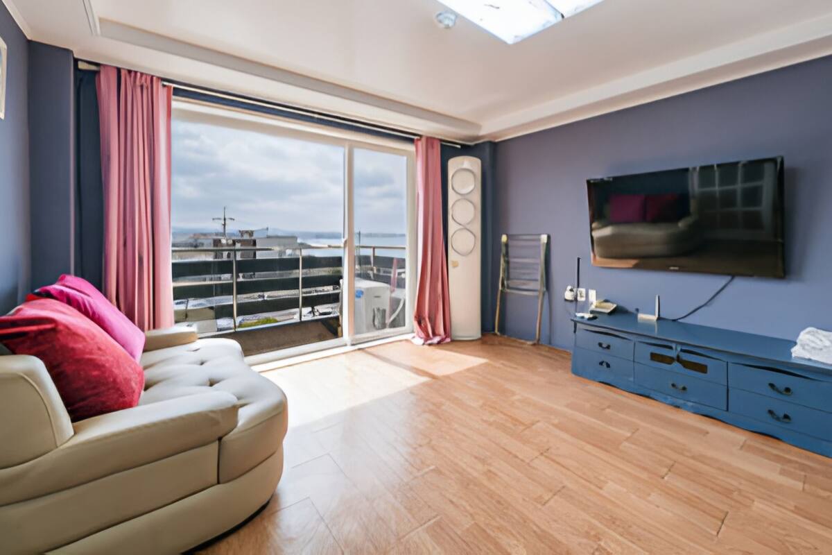 Ein modernes Wohnzimmer mit einer Couch, einem Flachbildfernseher, einem blauen Schrank und einer großen Glasschiebetür, die auf einen Balkon mit Blick auf die Stadt führt.
