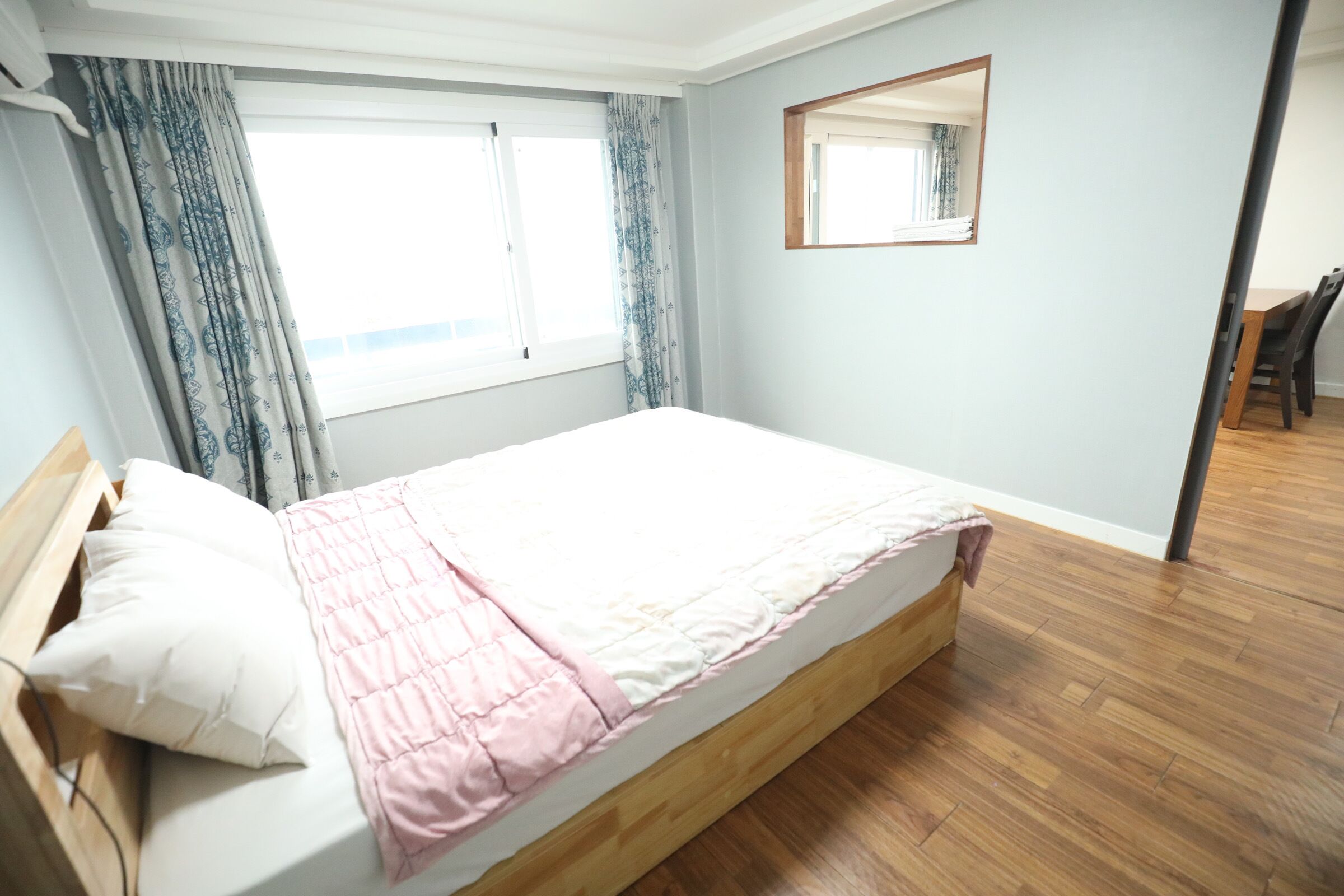 Ein Schlafzimmer mit Bett, Holz-Kopfteil, einem Fenster mit Vorhängen, einem Spiegel an der Wand und einem Essbereich im Hintergrund.