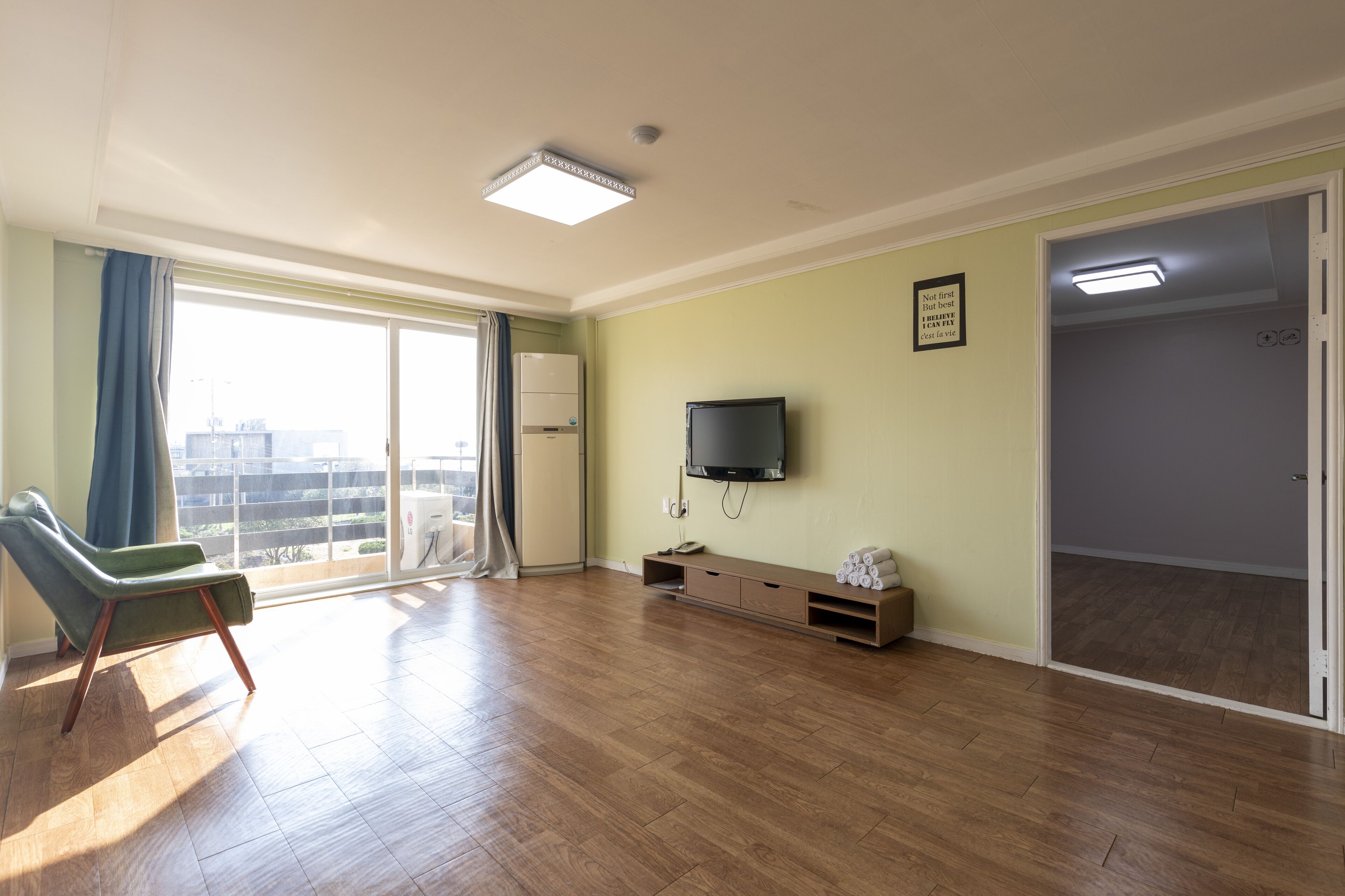 A spacious living room with a large sliding glass door leading to a balcony, a flat-screen TV mounted on the wall, and a wooden floor.