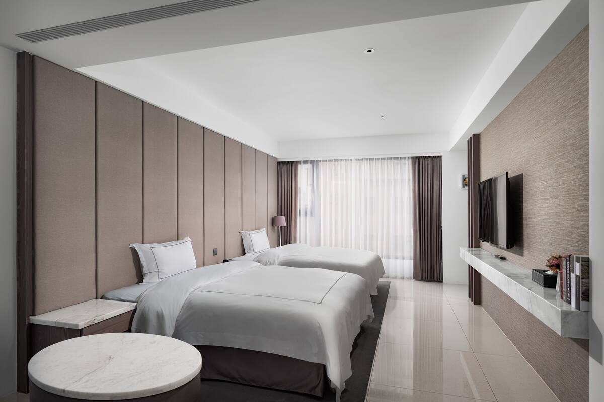 A modern hotel room with a large bed, a smaller bed, a seating area, and a TV mounted on the wall.