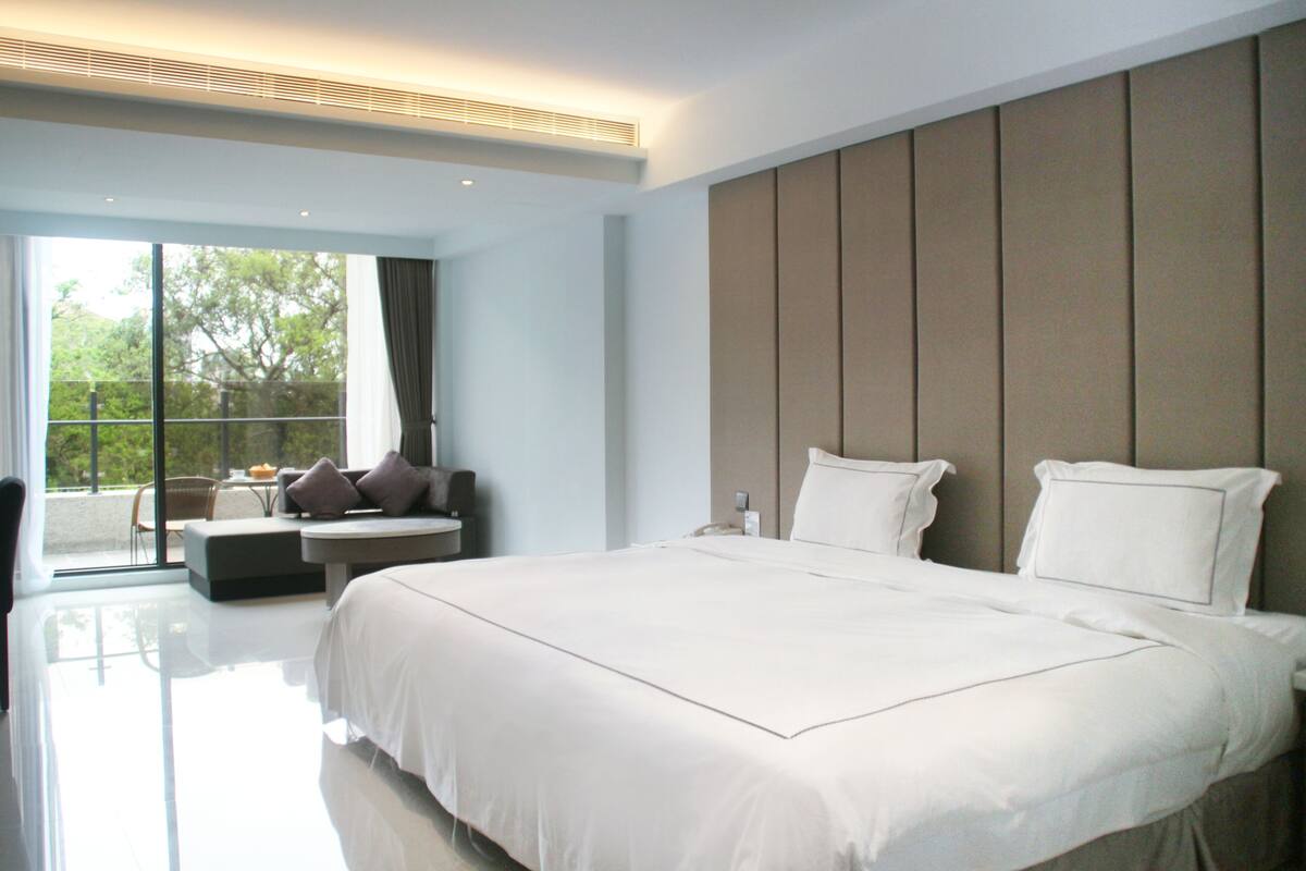A modern hotel room with a large bed, a seating area with a table, and a view of greenery outside.