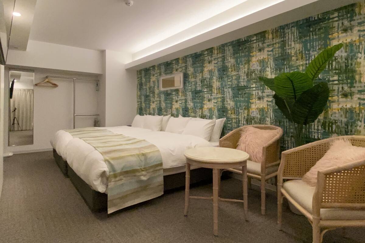 A modern hotel room with a large bed, two wicker chairs, a small round table, and a large green plant.