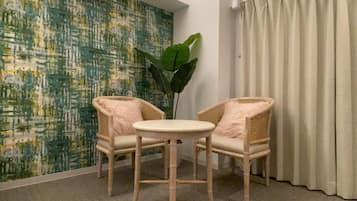 A room with two wicker chairs, a small round table, a large green plant, a textured wall, and a window with curtains.
