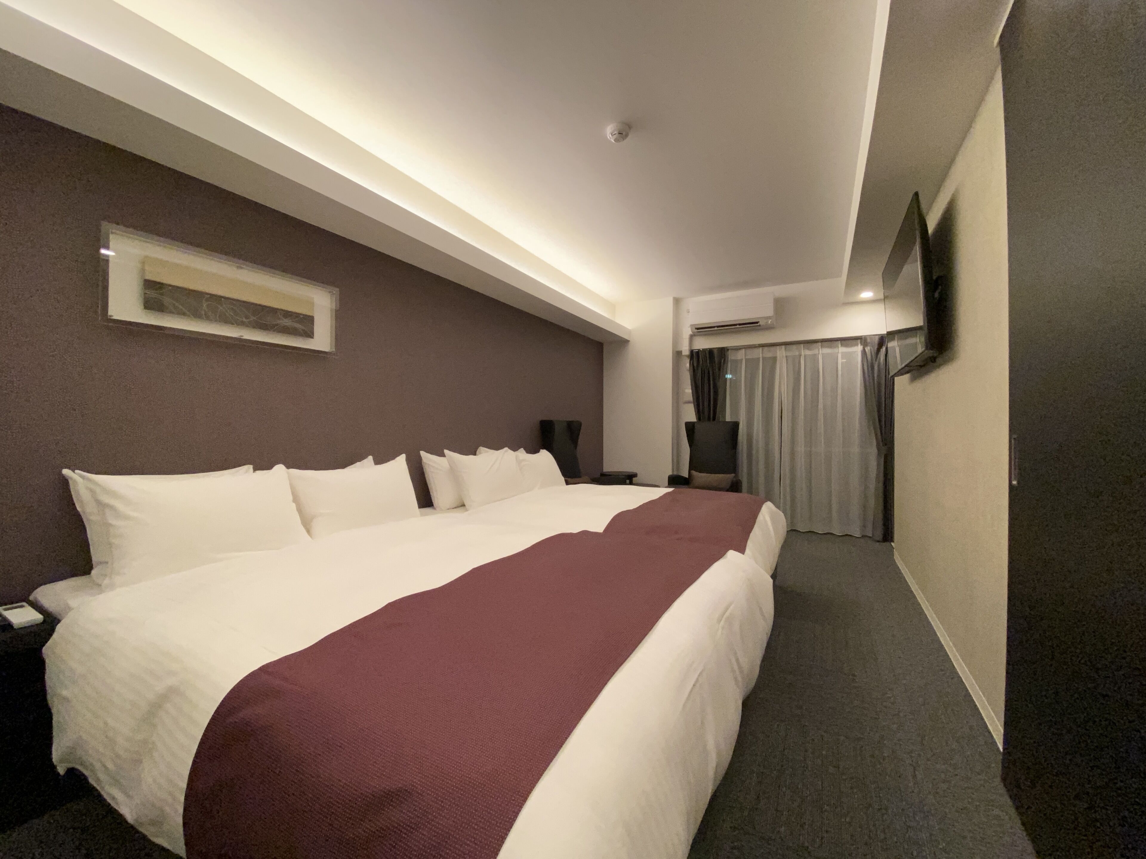 A hotel room with a large bed, a flat-screen TV, an air conditioning unit, and a window with curtains.