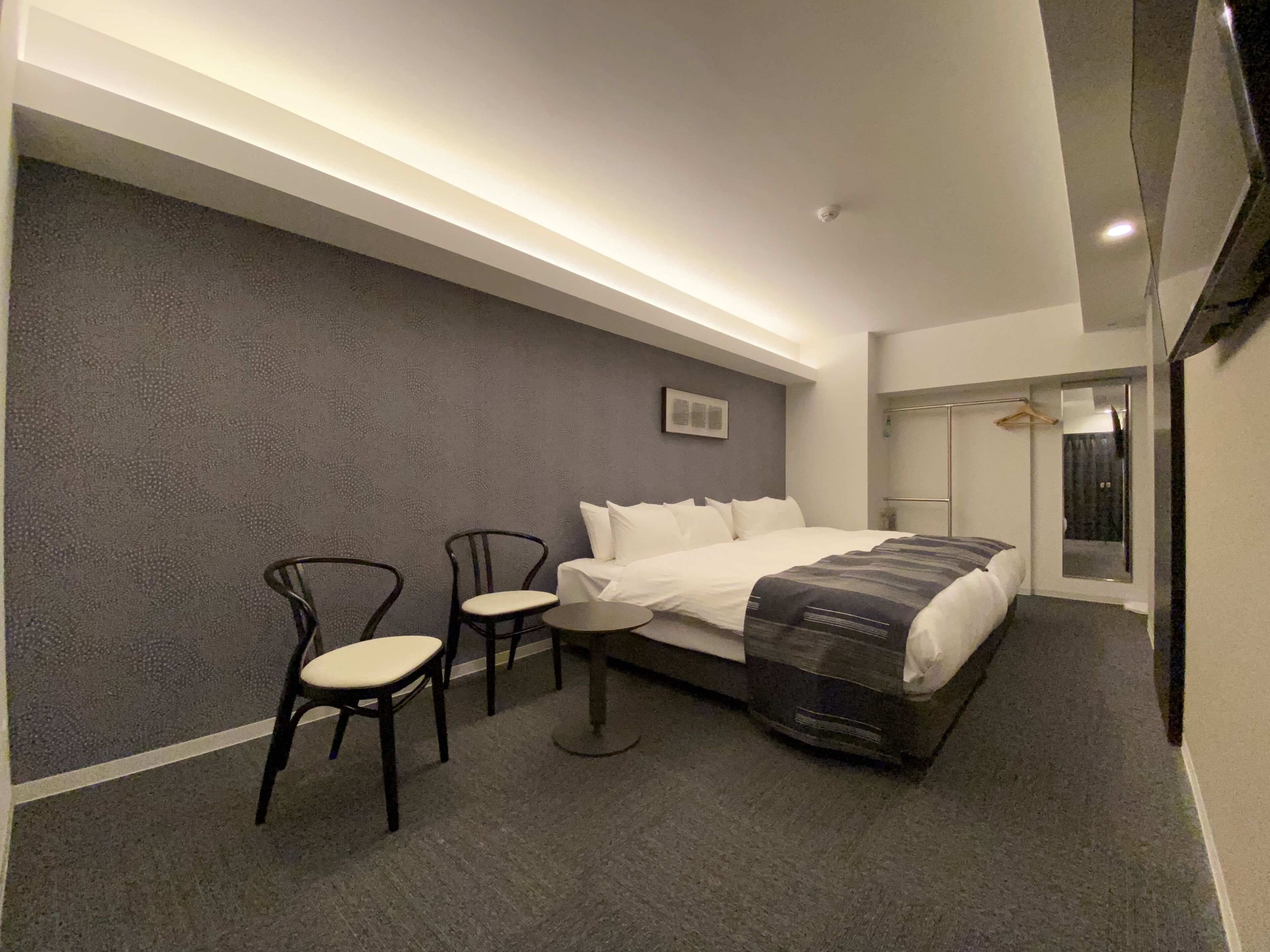 A modern hotel room with a large bed, two chairs, a small table, a mirror, and a picture on the wall.