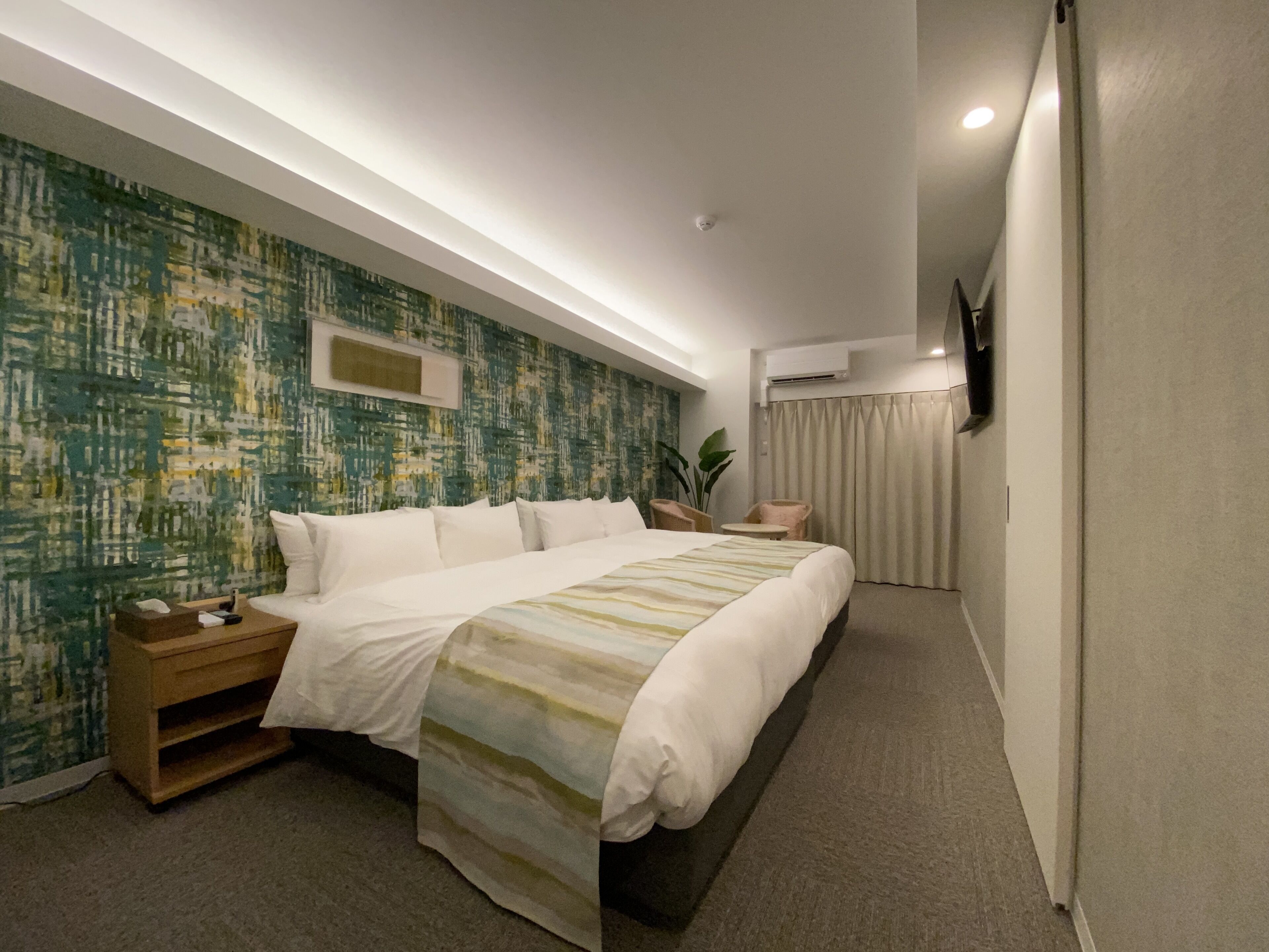 Een hotelkamer met een groot bed, een nachtkastje, een klein tafeltje, een potplant en een aan de muur bevestigde televisie.