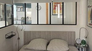 A modern bedroom with a large bed, a hanging light fixture, a bedside table with a lamp, and a mirror on the wall. - Charming 2-room apartment, city center, loggia, parking (Sète)