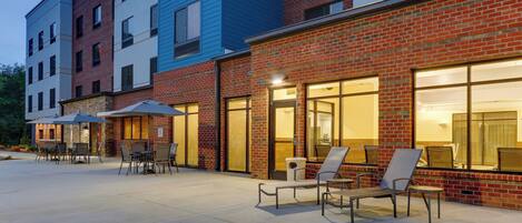 A building with a brick facade and large windows, outdoor seating area with chairs and umbrellas, and a paved patio.
