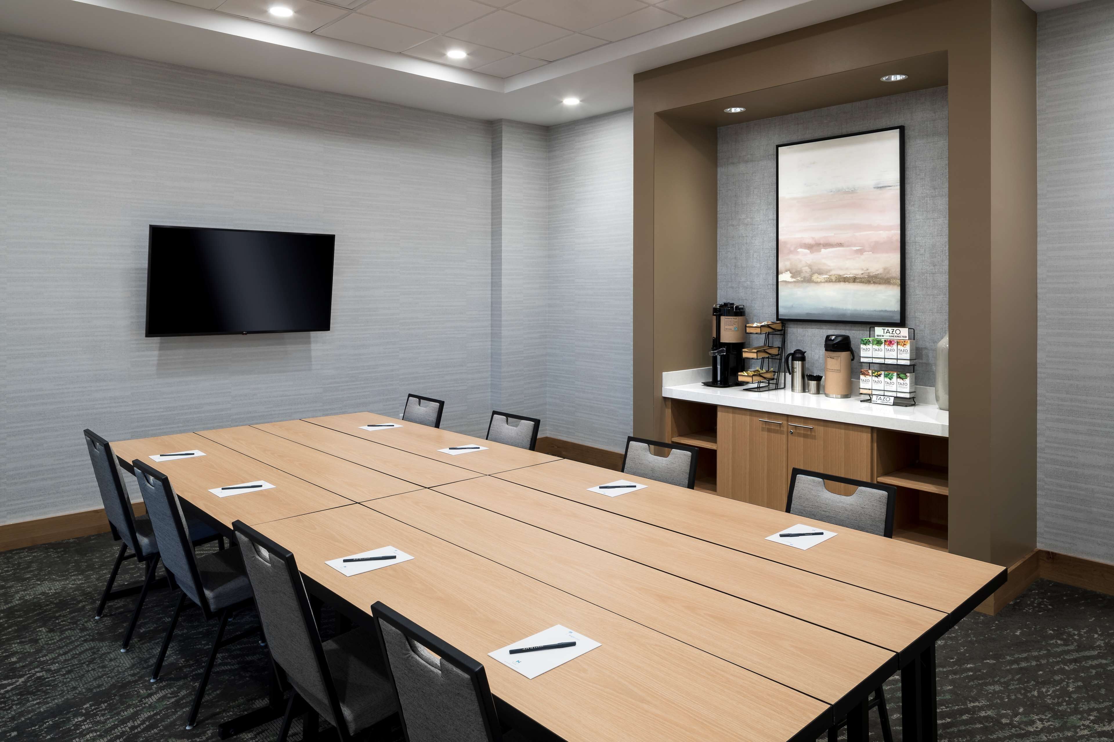 Een vergaderruimte met een lange tafel, stoelen, een flatscreen televisie, een koffiehoek en een schilderij aan de muur.