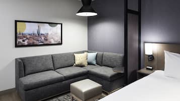 Ein modernes Hotelzimmer mit einer grauen Eckcouch, einem kleinen Hocker, einem Bett mit weißen Bettwäsche und einem Nachttisch mit Lampe, sowie einem an der Wand befestigten Kunstwerk, das eine Wüstenlandschaft zeigt.