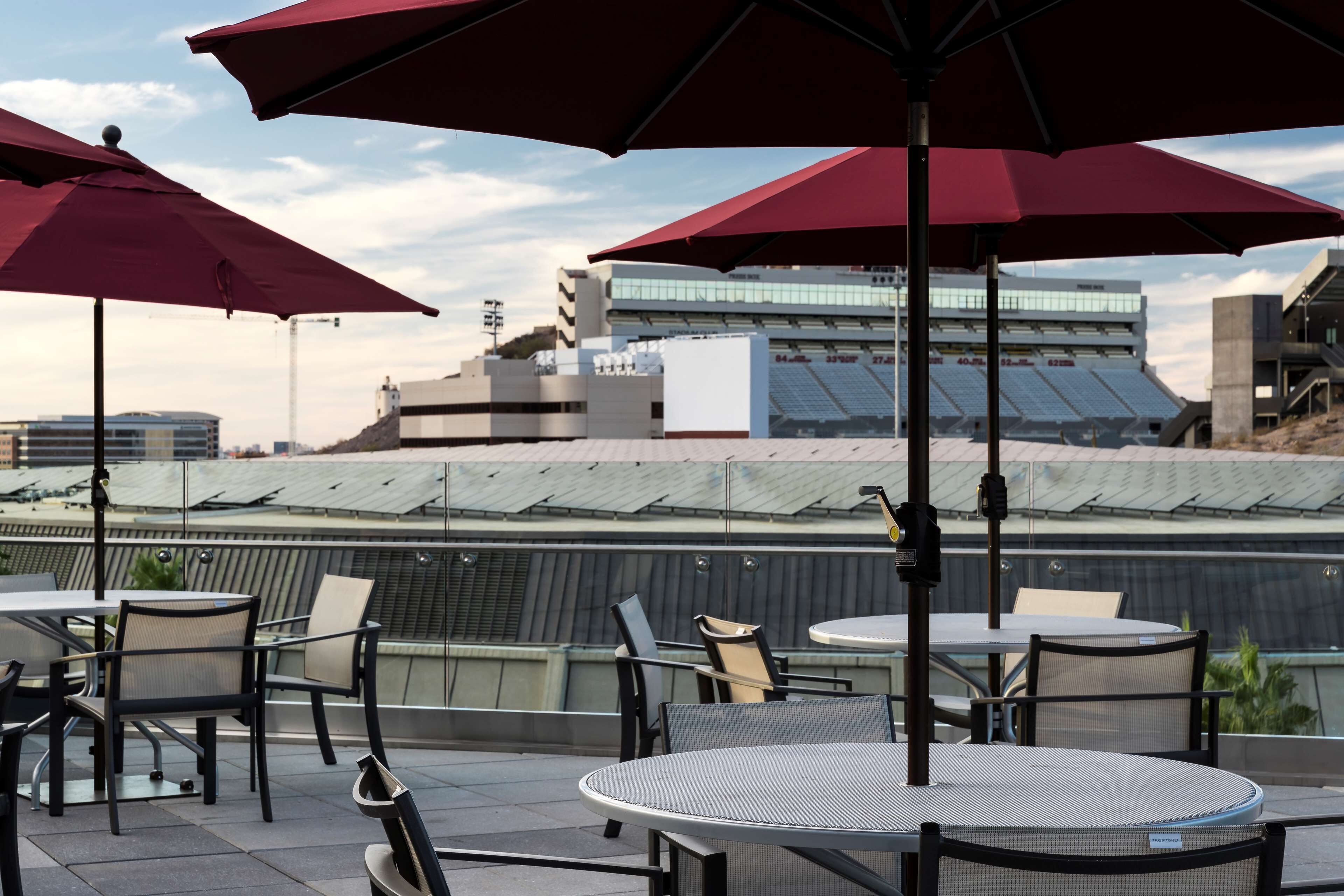 Outdoor seating with tables and chairs, red umbrellas, and a view of a stadium with a glass roof.