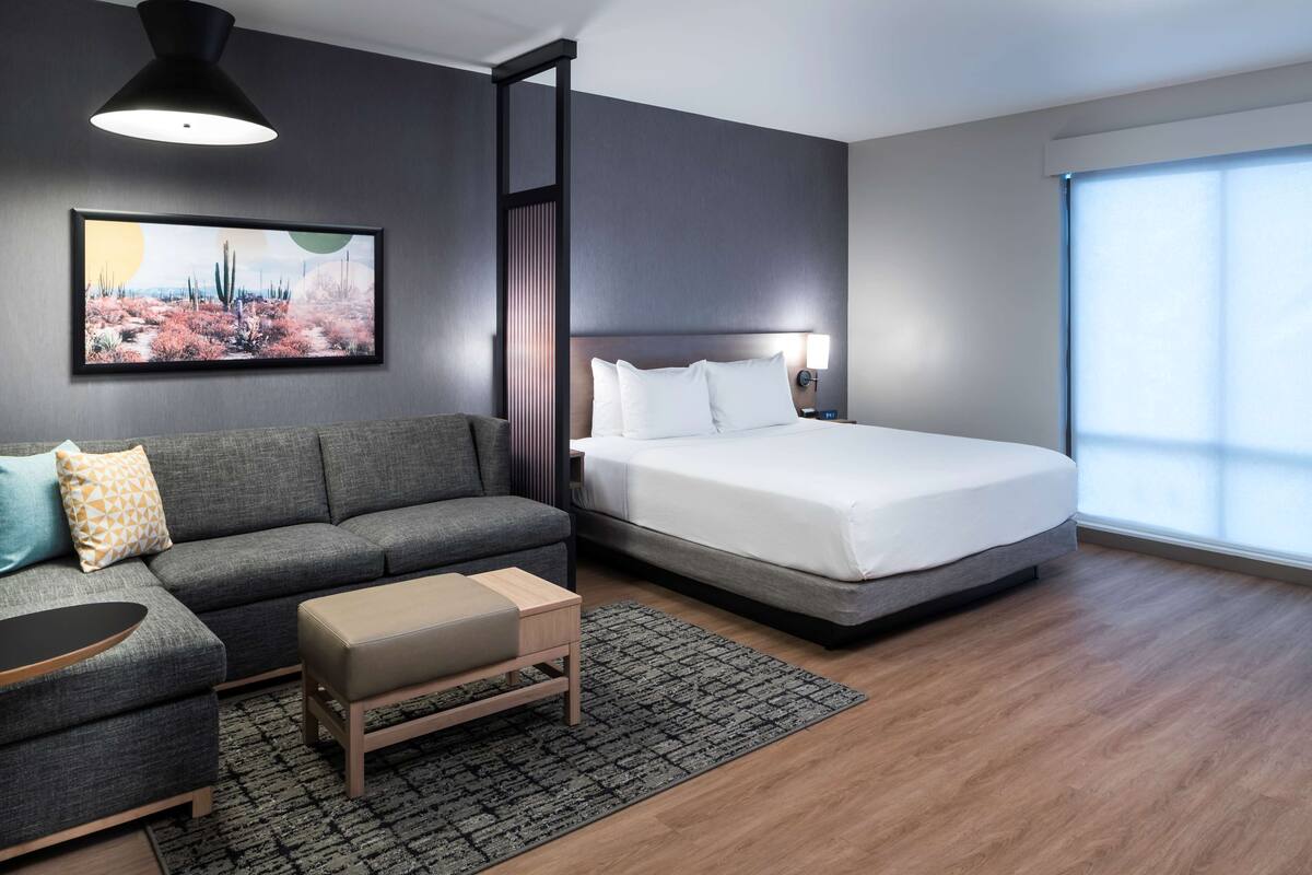 Moderni hotellihuone, jossa on harmaa sohva, sänky, pieni pöytä ja suuri ikkuna.