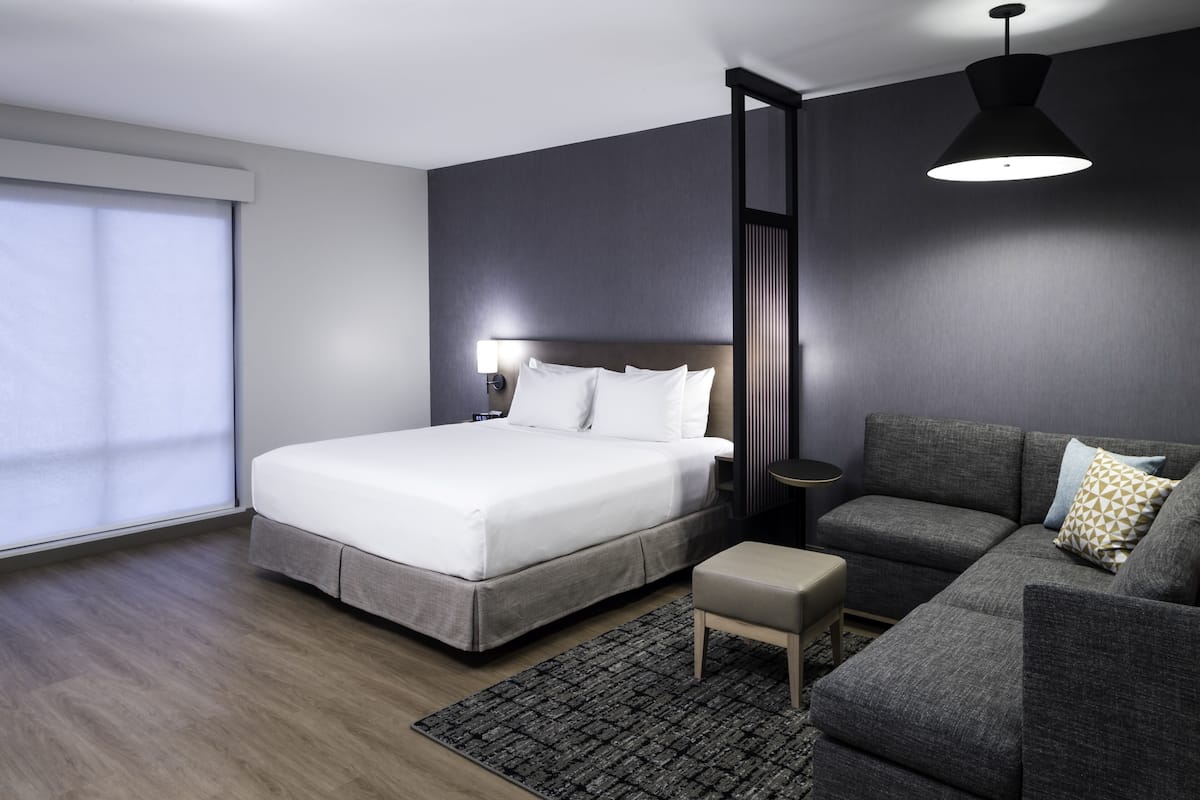 Moderni hotellihuone, jossa on suuri sänky, harmaa sohva, pieni pöytä ja suuri ikkuna, jossa on sälekaihtimet.