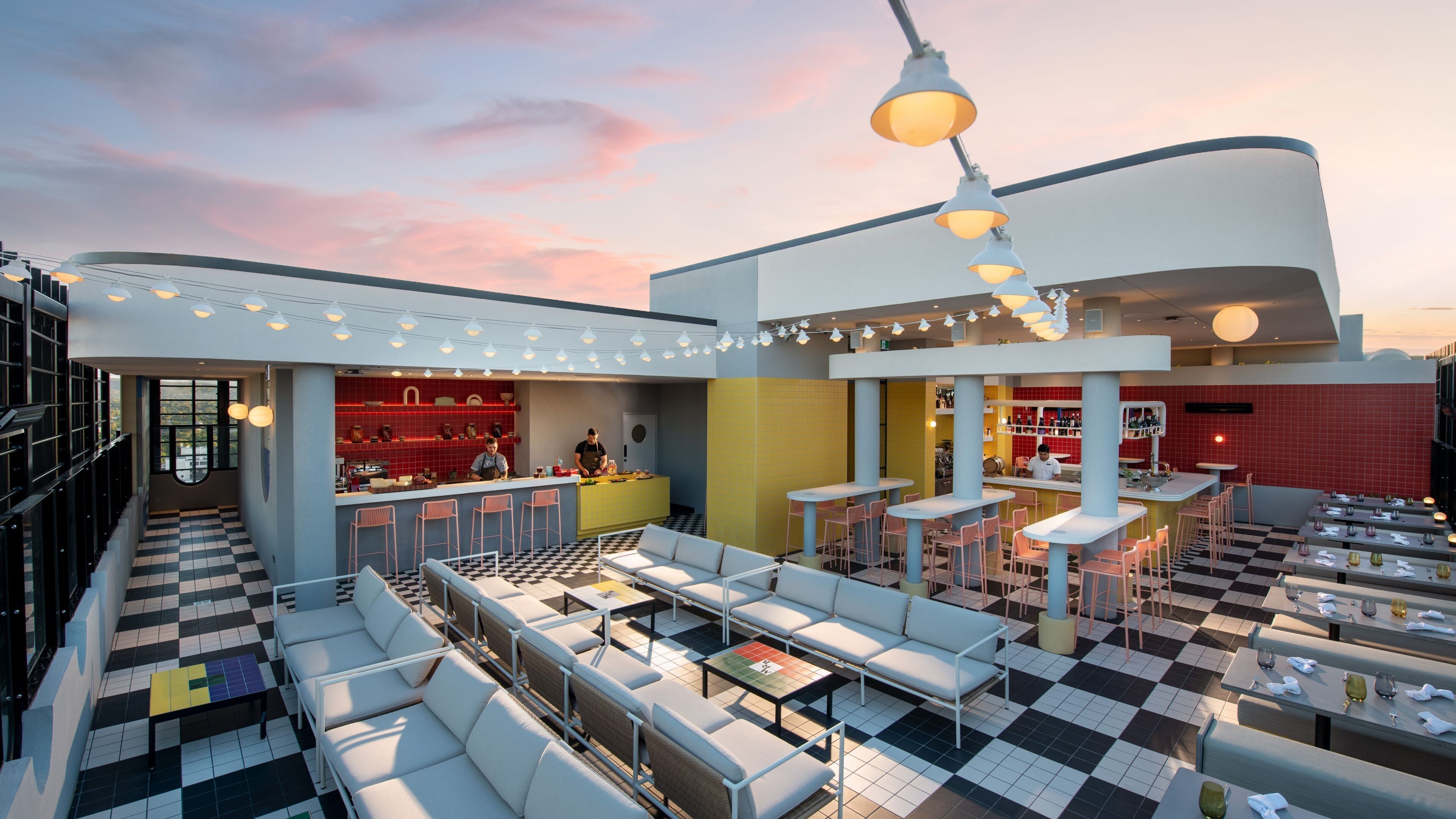 Un bar situé sur un toit, avec un sol à carreaux, des places assises en plein air et un comptoir agrémenté d’un mur recouvert de carreaux rouges.