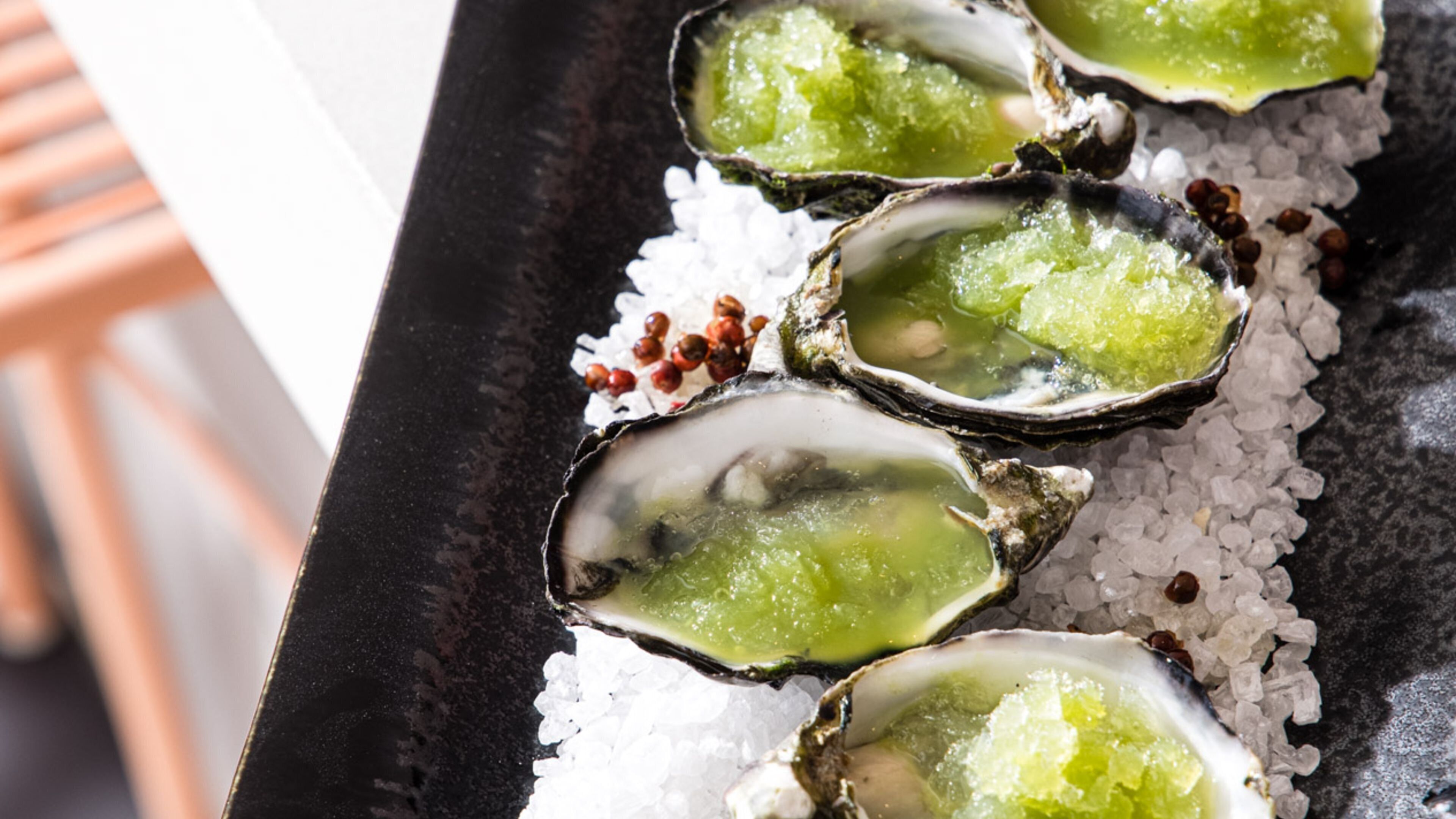 A tray of oysters on ice with a green topping, possibly a sauce or garnish, and some red peppercorns.