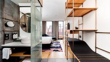 Une chambre d’hôtel moderne dotée d’une paroi en verre, d’un grand lit, d’un bureau intégré, d’une salle de bain avec baignoire et offrant une vue sur la ville.