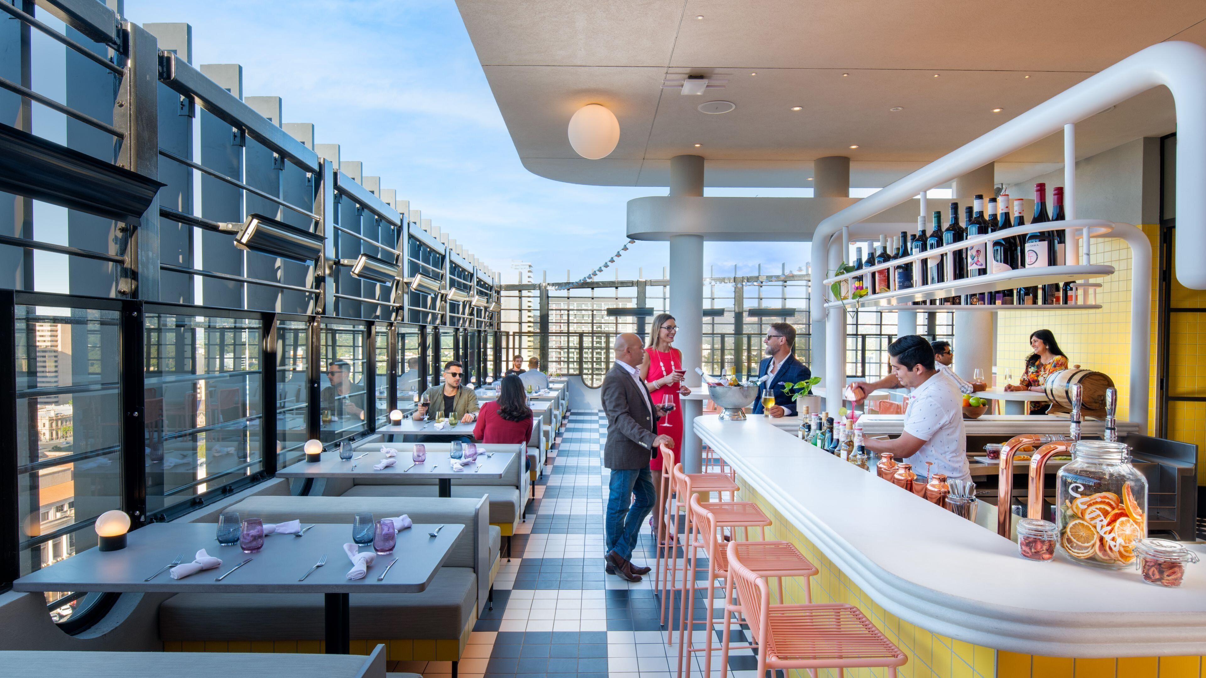 An outdoor dining area with tables and chairs, a bar with bottles and a bartender, and a city view through large windows.