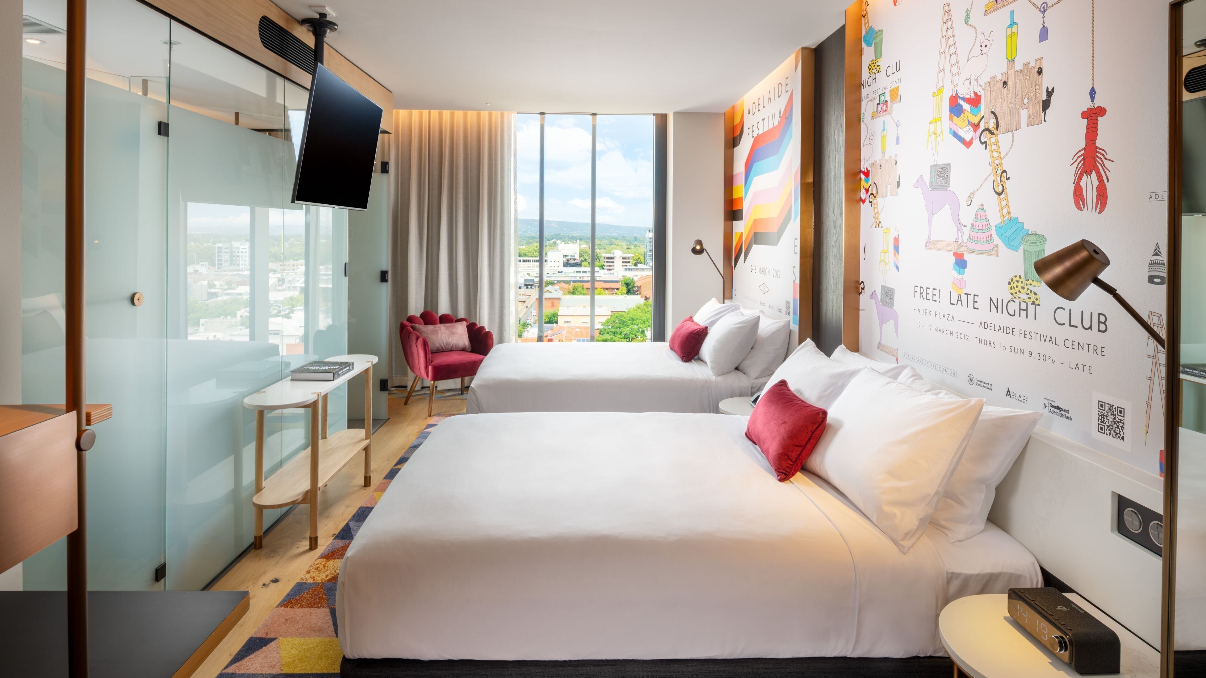 A hotel room with two beds, a red chair, a small table, a TV, and a large window with a city view.