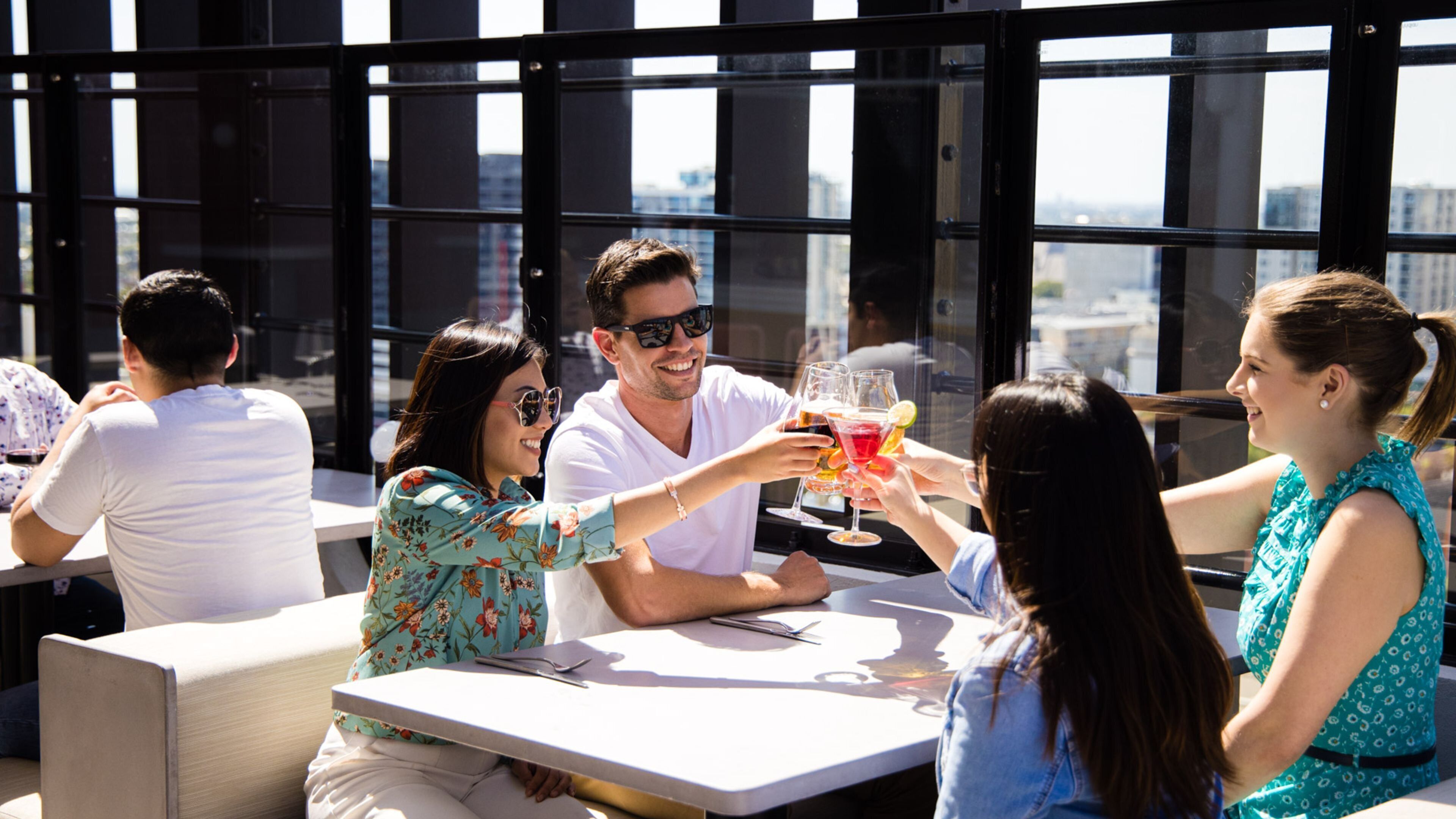 A group of people toasting with drinks at an outdoor table with a city view.
