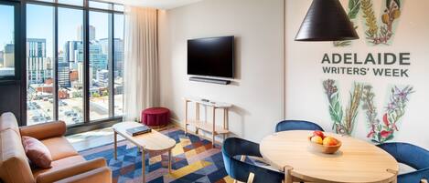 A modern hotel room with a large window, a flat-screen TV, a round wooden table with chairs, a red ottoman, and a colorful geometric carpet.