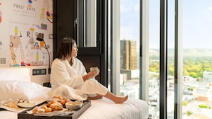 A person sitting on a bed in a room with a city view, holding a cup. There is a tray with food items on the bed. - Hotel Indigo Adelaide Markets by IHG (Adelaide)