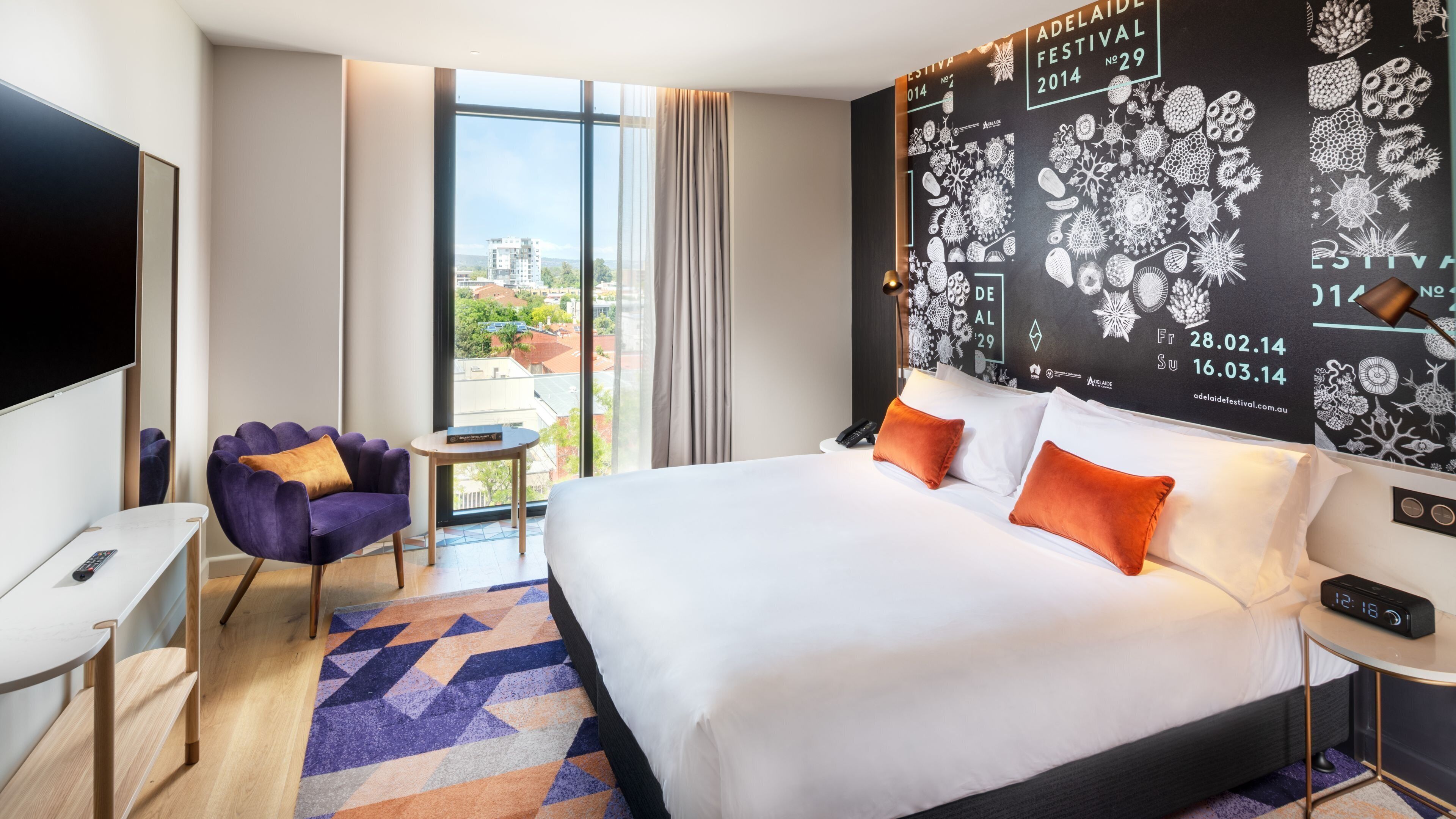 A hotel room with a large bed, a TV, a small table, a chair, and a view of the city.