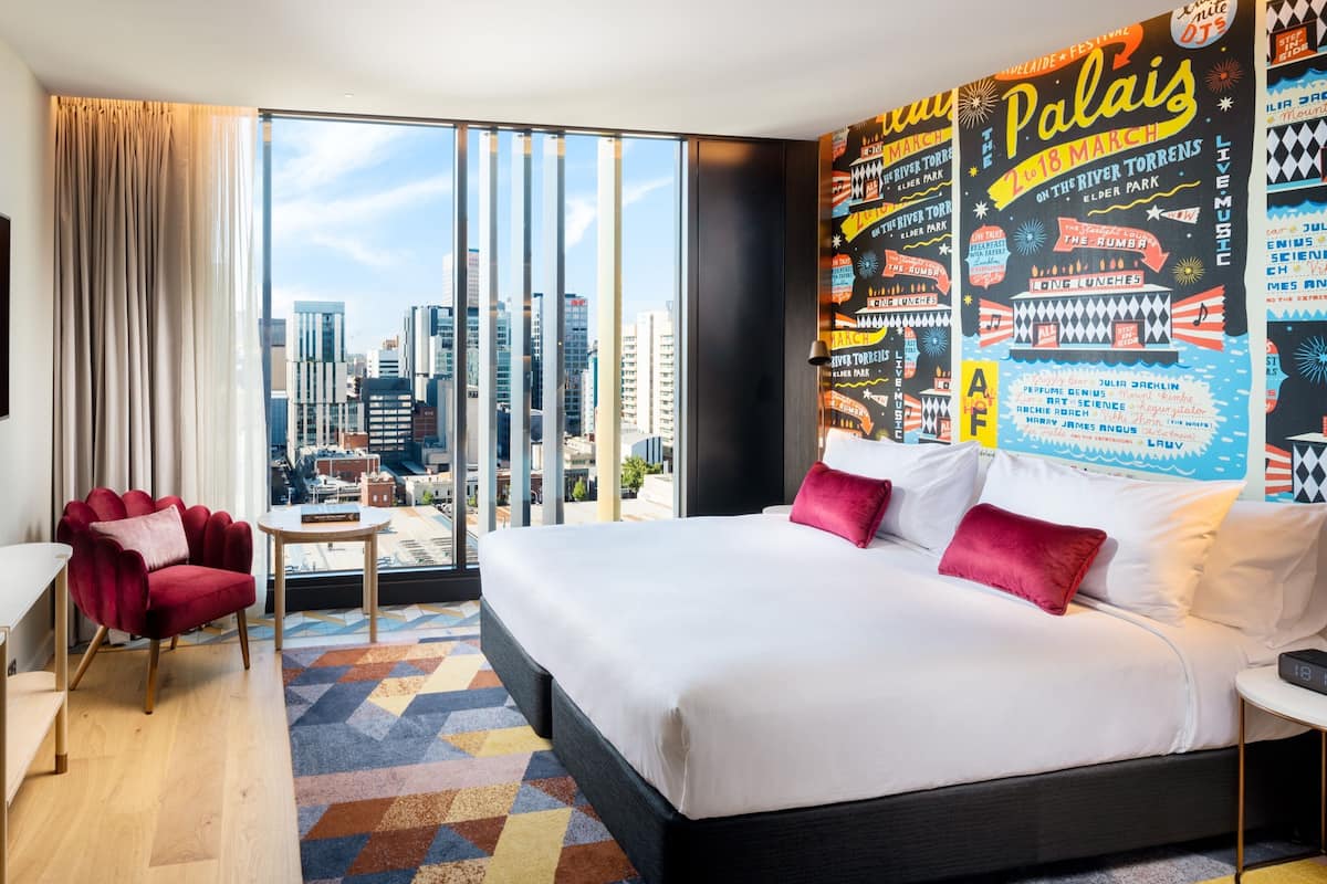 A modern hotel room with a large bed, a red armchair, a small table, and a city view through floor-to-ceiling windows.