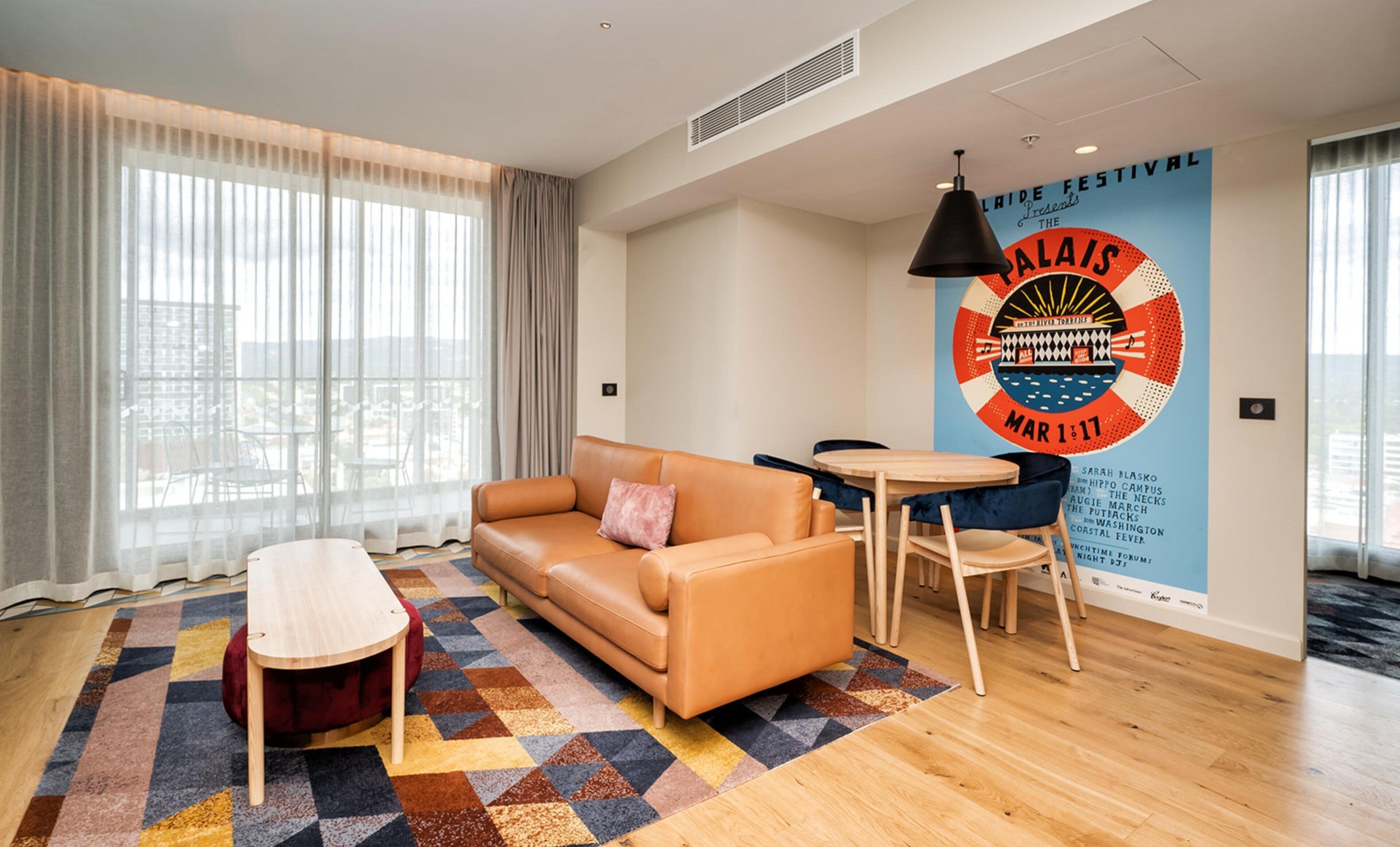 A modern hotel room with a sofa, a small table, and a chair. There is a large window with sheer curtains, a patterned rug, and a wall with a poster advertising an event.